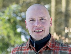 Bildbeschreibung: Kopfbild von Frank Schmidt, Hintergrund Wald, er hat einen Pullower und darüber ein kariertes Hemd an