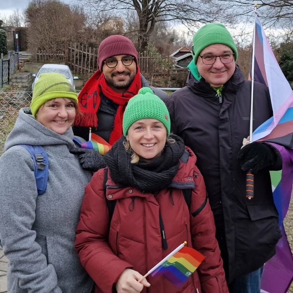 Bildbeschreibung; Gruppenbild: Antje Naumann, Kassem Taher Saleh (MdB), Berit Koch und Jens Bitzka (v.l.n.r) , Antje und Berit halten jeweils eine kleine Fahne in Regenbogenfarben, Jens hält eine große Fahne in Regenbogenfarben, Antje, Berit und Jens haben Grüne Mützen auf, Kassem trägt eine rote Mütze und einen roten Schal