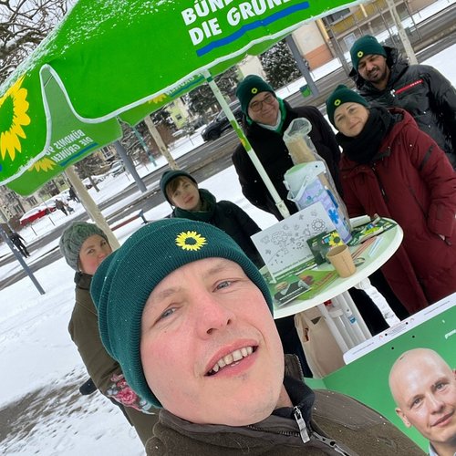 Bildbeschreibung: Selfie von Frank Schmidt (er trägt eine grüne Mütze mit kleinem Sonnenblumensymbol. Im Hintergrund: Stehtisch mit grünem Schirm, Teil von seinem Wahlplakat, am Tisch stehen 5 Personen, 3 davon mit einer grünen Mütze mit Sonnenblumensymbol, im Hintergrund eine Straße und ein Haus, auf dem Boden liegt Schnee 