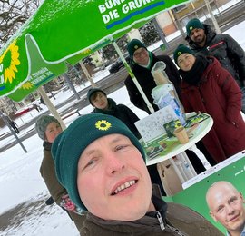 Bildbeschreibung: Selfie von Frank Schmidt (er trägt eine grüne Mütze mit kleinem Sonnenblumensymbol. Im Hintergrund: Stehtisch mit grünem Schirm, Teil von seinem Wahlplakat, am Tisch stehen 5 Personen, 3 davon mit einer grünen Mütze mit Sonnenblumensymbol, im Hintergrund eine Straße und ein Haus, auf dem Boden liegt Schnee 