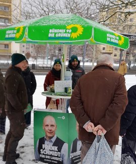 Bildbeschreibung: 7 Personen stehen um den Stehtisch (Infotisch) mit dem Grünen Parteisonnenschirm, darunter Bundestagskandidat Frank Schmidt, im Hintergrund eine Straße und Hochhäuser