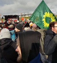 Bildbeschreibung: Blick Richtung Bühne der Hauptkundgebung, dazwischen viele Menschen, im Vordergrund eine Grüne Fahne mit Sonnenblumensymbol (Parteifahne B 90/GRÜNE)