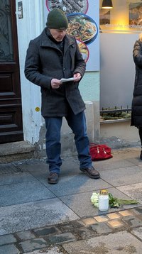 Frank Schmidt, steht nehmen den Stolpersteinen und hält eine Rede, er schaut auf ein Papierblatt. er hat eine blaue Hose und eine graue Jacke an, er Trägt eine grüne Stickmütze.