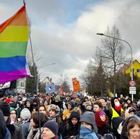 Bildbeschreibung: Viele Menschen auf dem Weg zum Kundgebungsort. Im Vordergrunf ist eine große Regenbogenfahne zu sehen. 